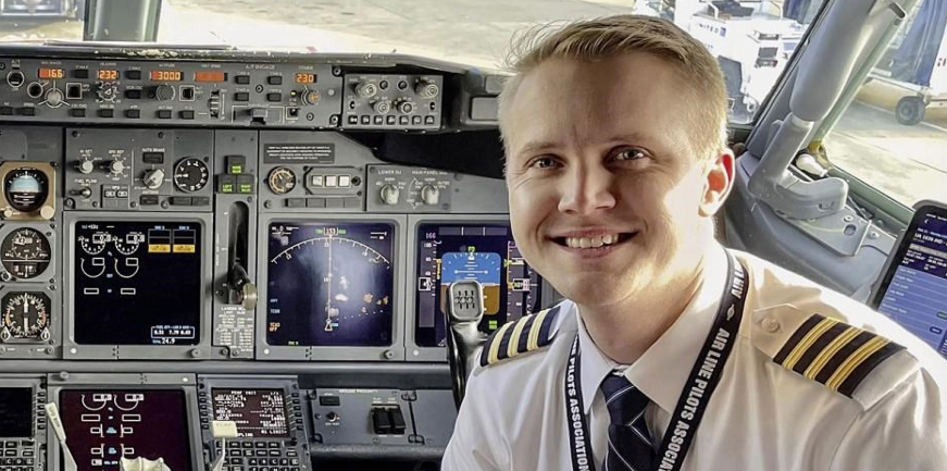 Lächelnder Pilot in Uniform im Cockpit, Instrumente und Steuerhörner sichtbar.